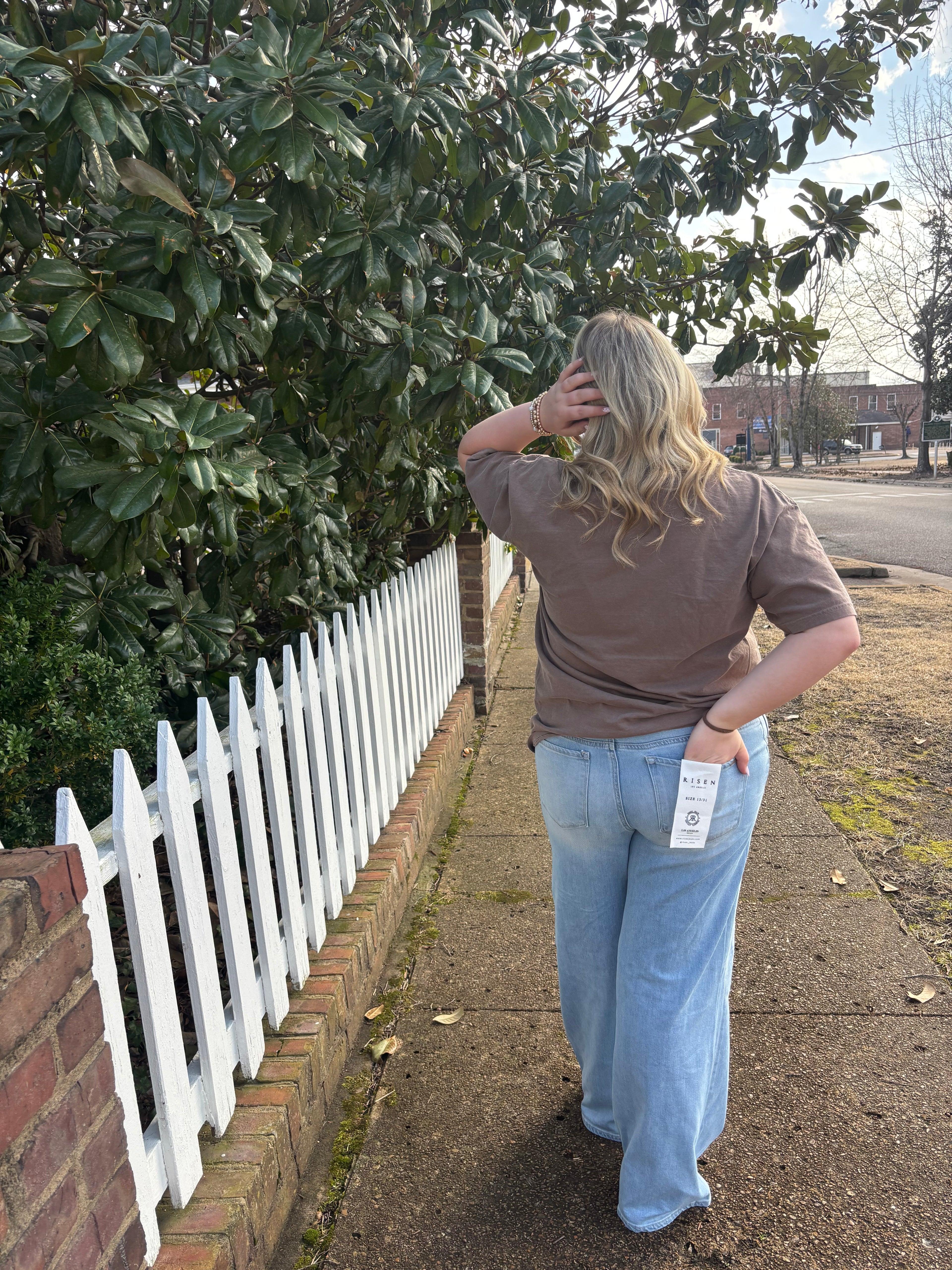A woman with her back turned towards the camera wearing light washed jeans with a brown t-shirt standing outside next to a white picket fence 