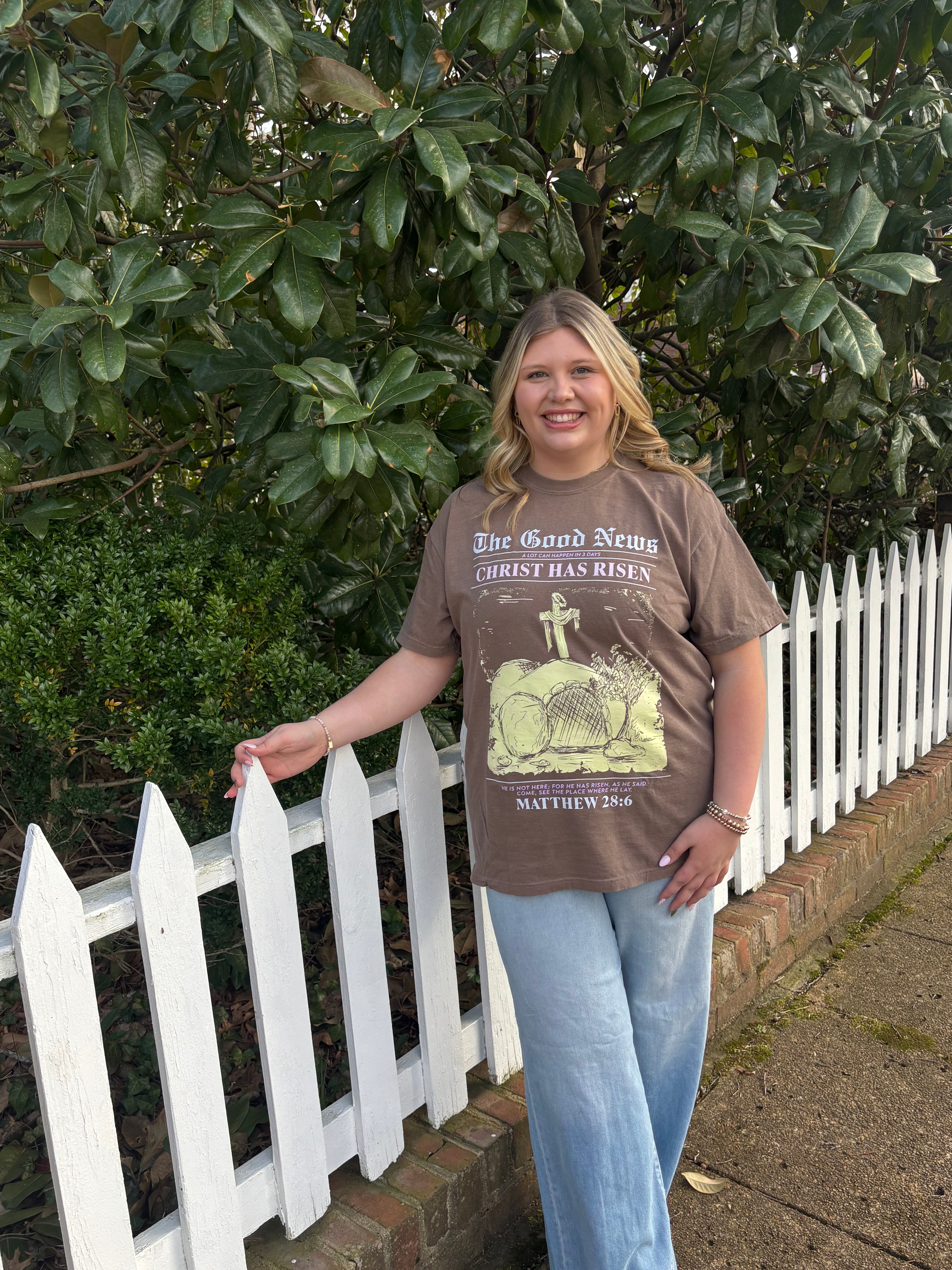A woman wearing light washed jeans with a brown t-shirt with faith based text on it standing outside next to a white picket fence with greenery behind her