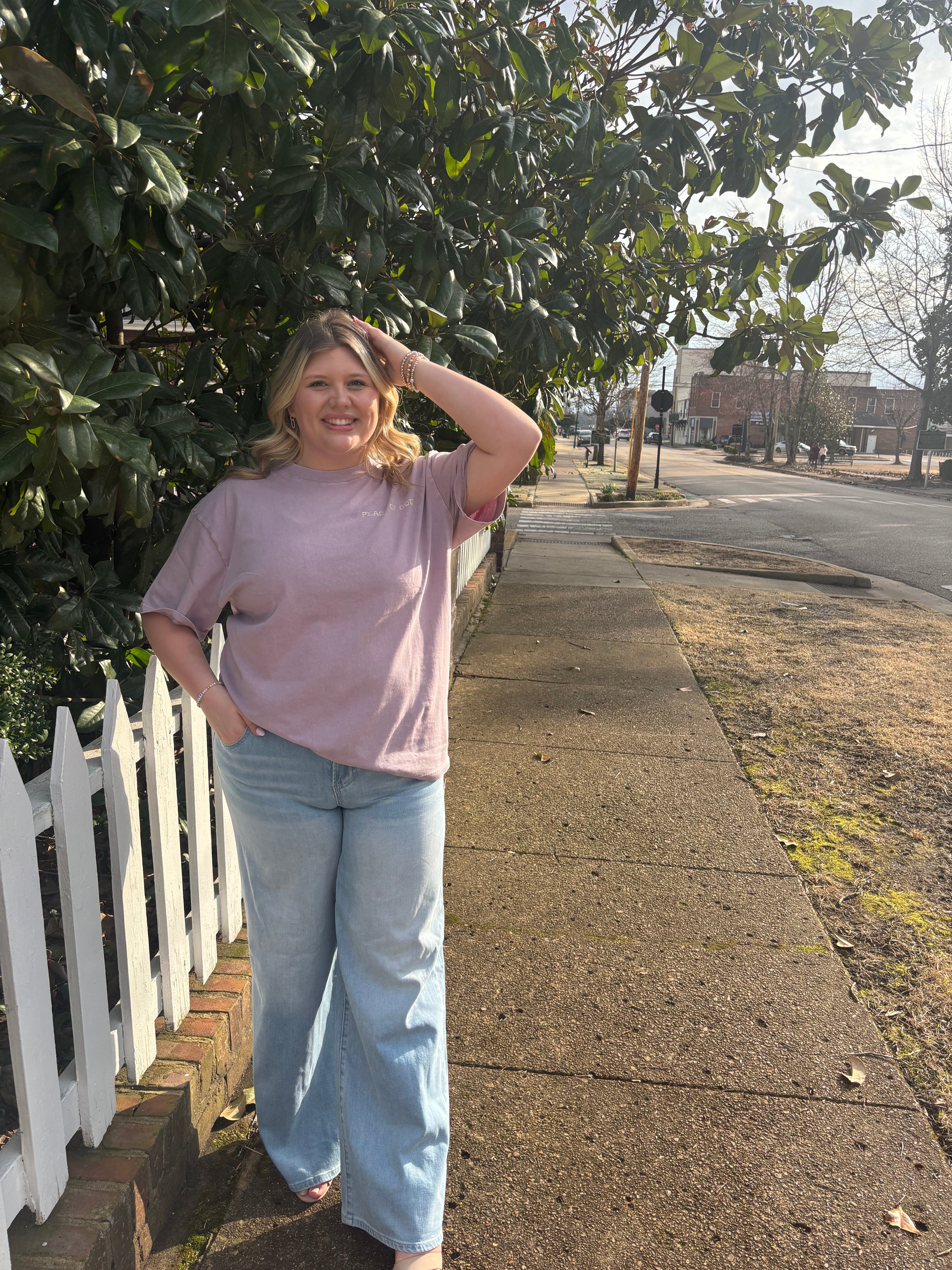 Person standing on a sidewalk next to a bush with a white fence in the background
