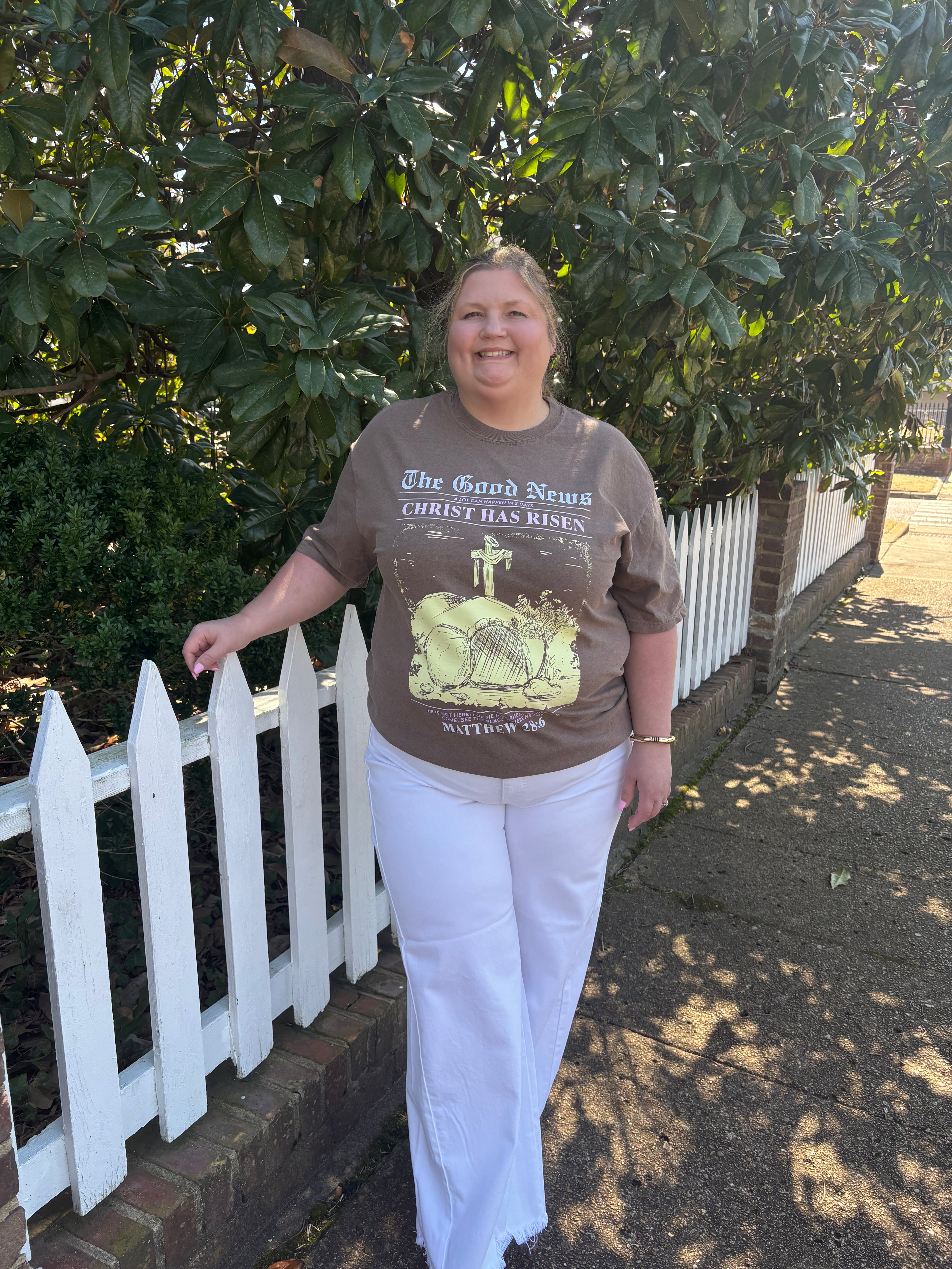 Person wearing a brown t-shirt with graphic design standing in front of a white picket fence.