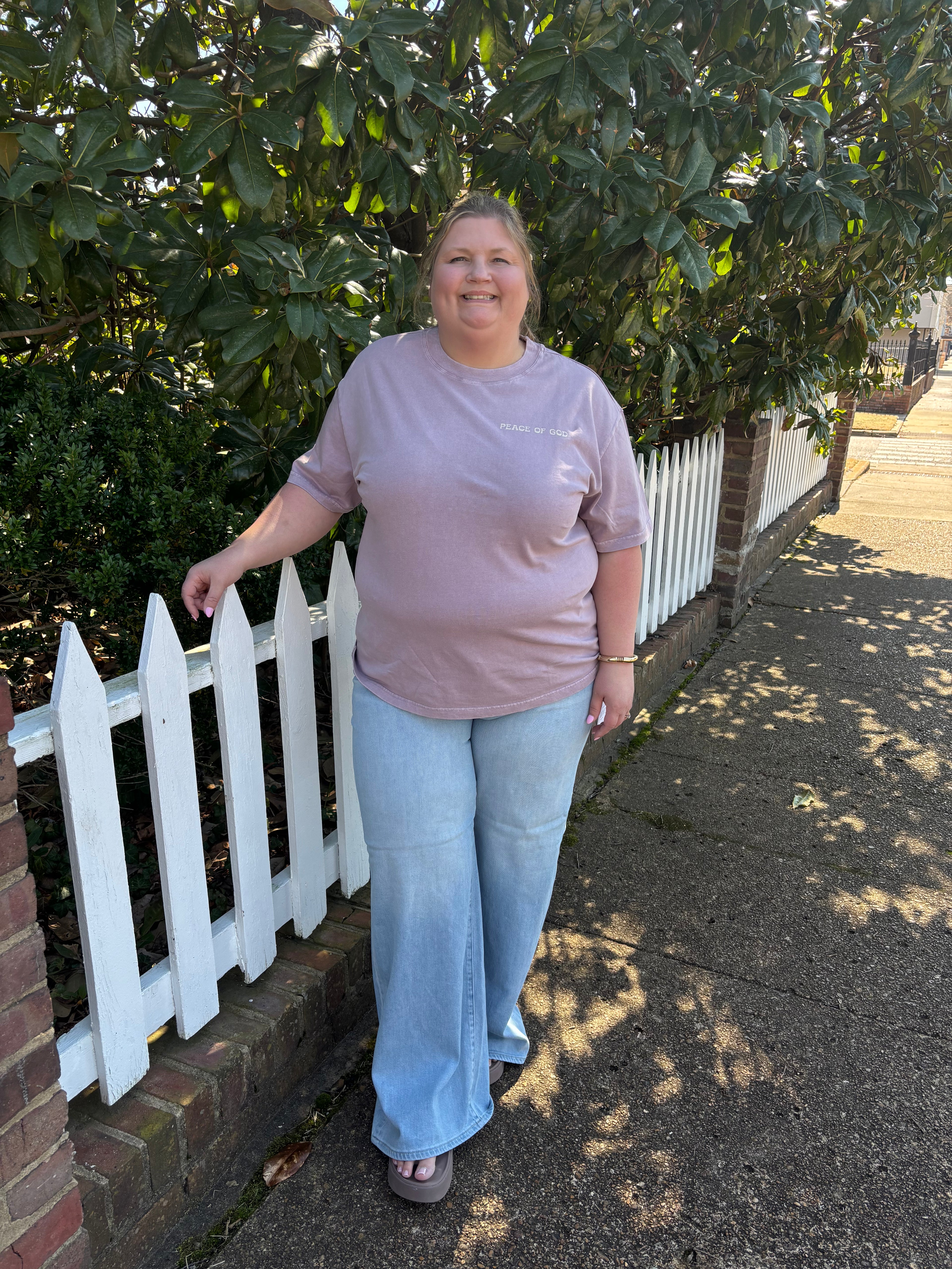 Woman standing on a sidewalk next to a white picket fence with greenery in the background