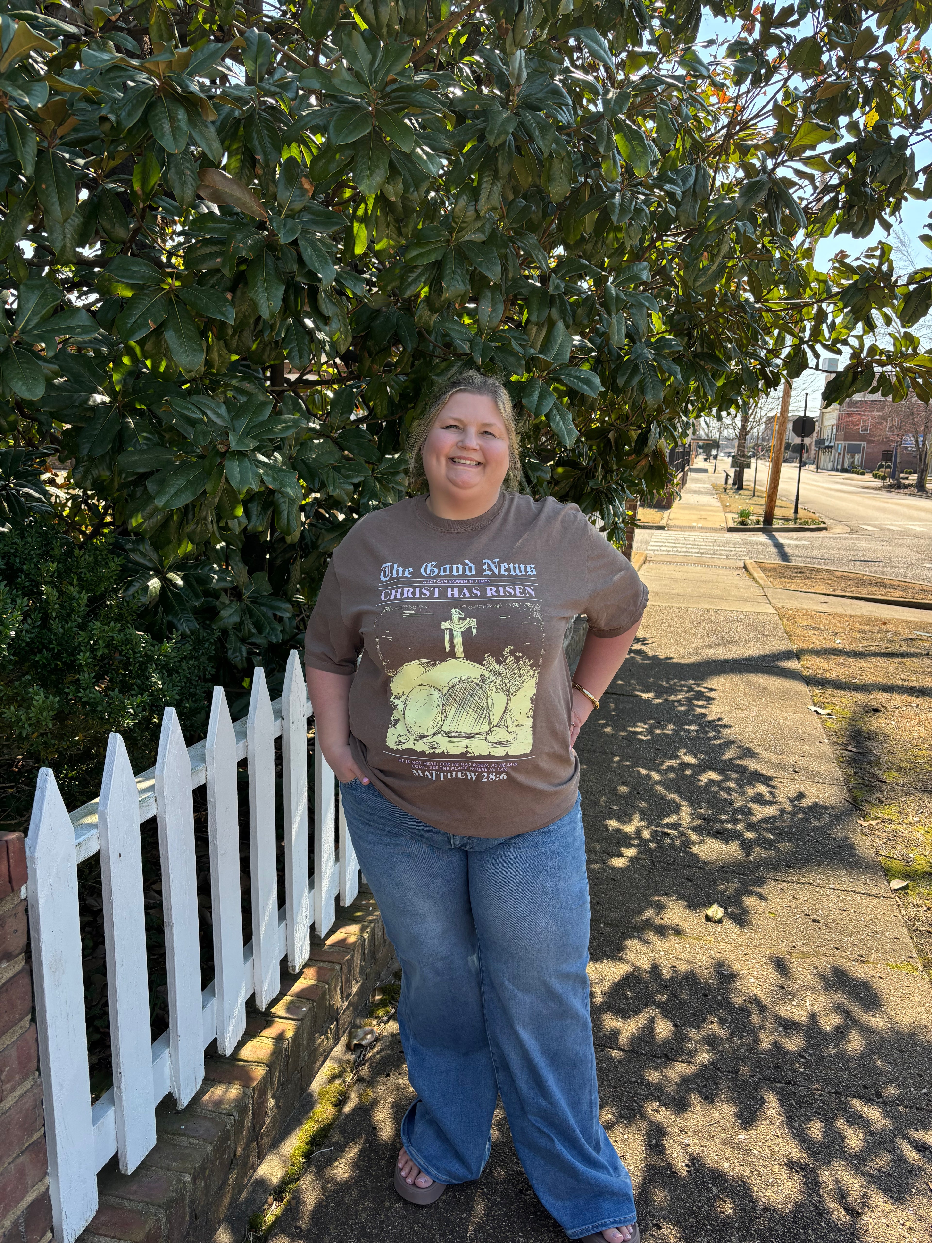 Woman wearing a graphic t-shirt standing in front of a white picket fence with trees in the background.