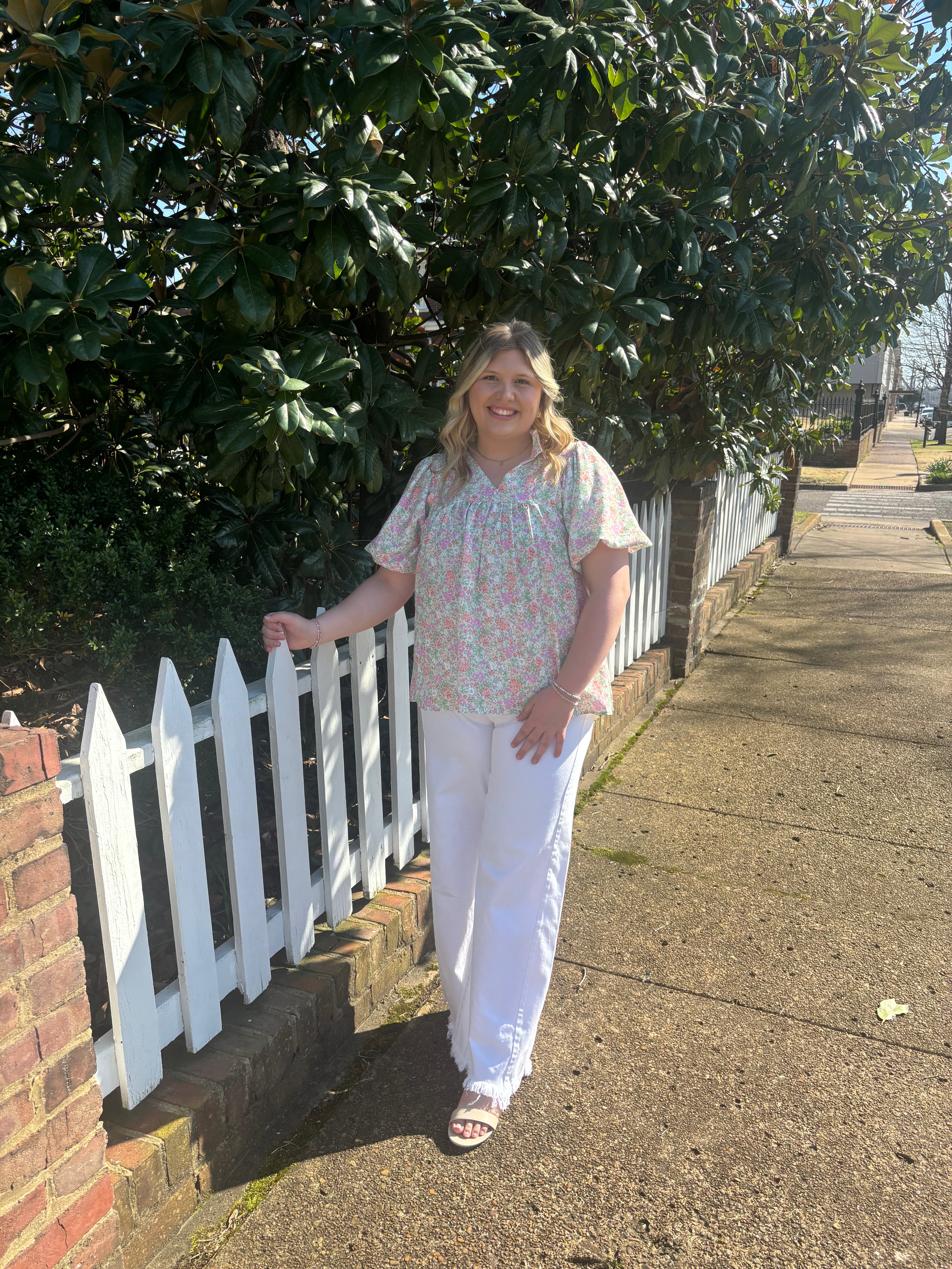 Woman standing on a sidewalk next to a white picket fence with greenery in the background