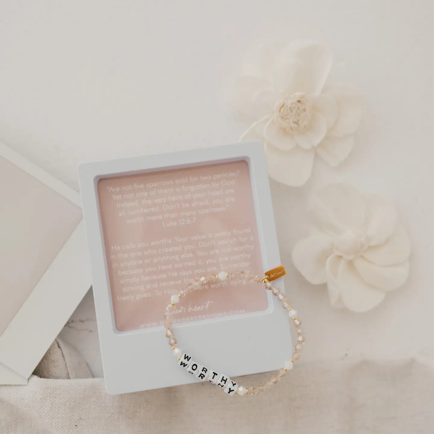 Bracelet with 'WORTHY' charm on a pink card with text, surrounded by flowers.